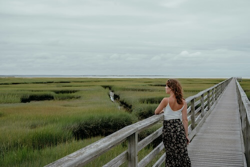 Woman leaning on a wooden boardwalk railing, looking out over a green salt marsh in a quiet moment of reflection and self-respect