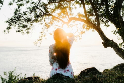 Woman with long dark hair sitting under a tree on a grassy slope, looking out at the sea at sunrise in a quiet moment of self-respect